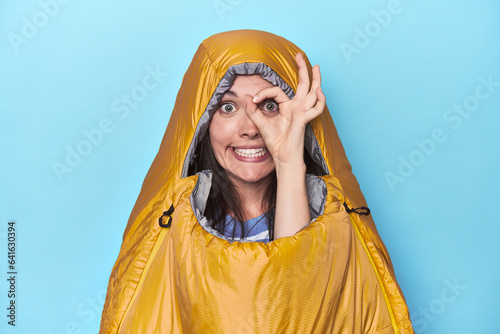 Woman in sleeping bag on blue background excited keeping ok gesture on eye.
