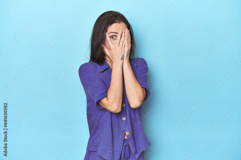 Young caucasian woman on blue backdrop blink through fingers frightened and nervous. Young caucasian woman on blue backdrop blink through fingers frightened and nervous.