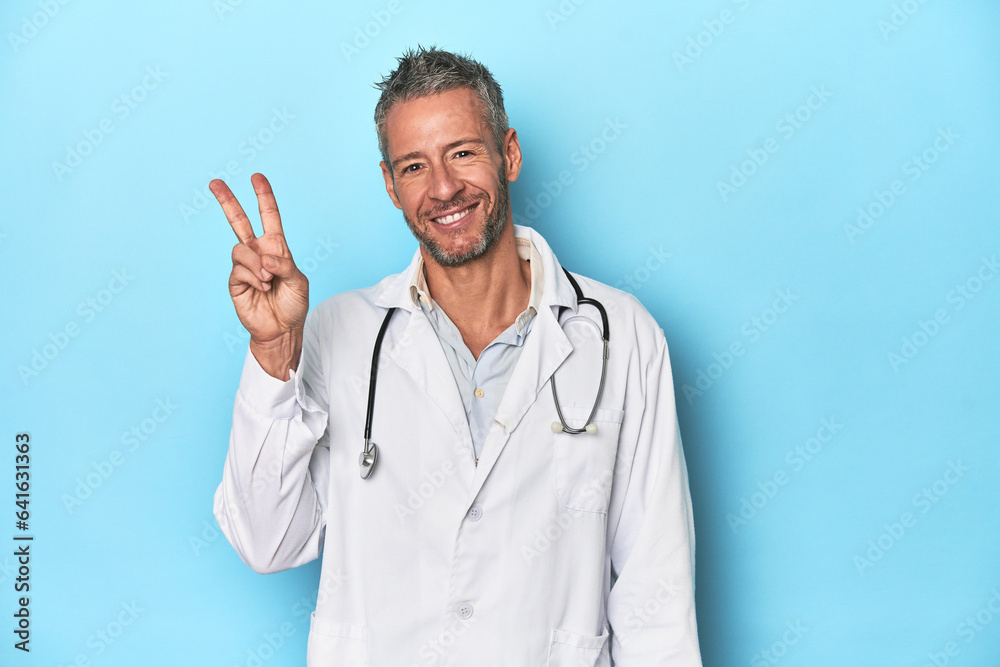 Caucasian middle-aged doctor on blue background joyful and carefree showing a peace symbol with fingers. Caucasian middle-aged doctor on blue background joyful and carefree showing a peace symbol with fingers.