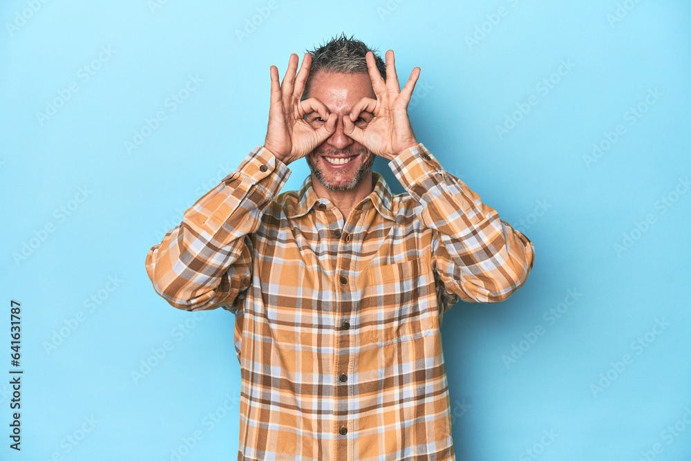 Middle-aged caucasian man on blue backdrop showing okay sign over eyes Middle-aged caucasian man on blue backdrop showing okay sign over eyes