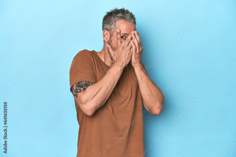 Middle-aged caucasian man on blue backdrop blink through fingers frightened and nervous. Middle-aged caucasian man on blue backdrop blink through fingers frightened and nervous.