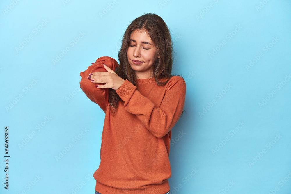 Teen girl in vibrant orange sweater on blue massaging elbow, suffering after a bad movement.