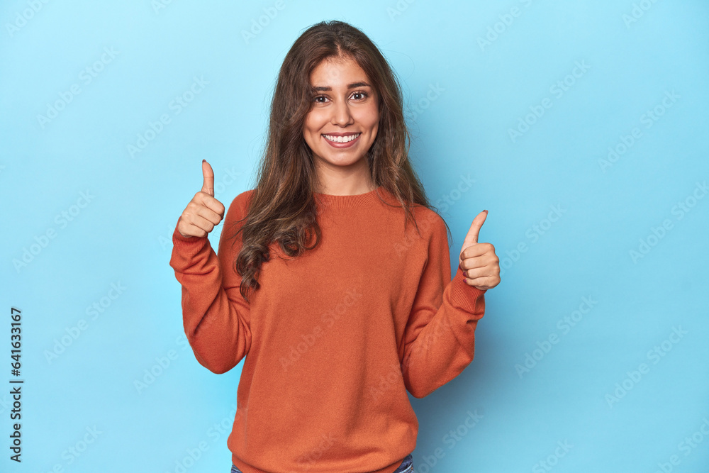 Teen girl in vibrant orange sweater on blue raising both thumbs up, smiling and confident. Teen girl in vibrant orange sweater on blue raising both thumbs up, smiling and confident.