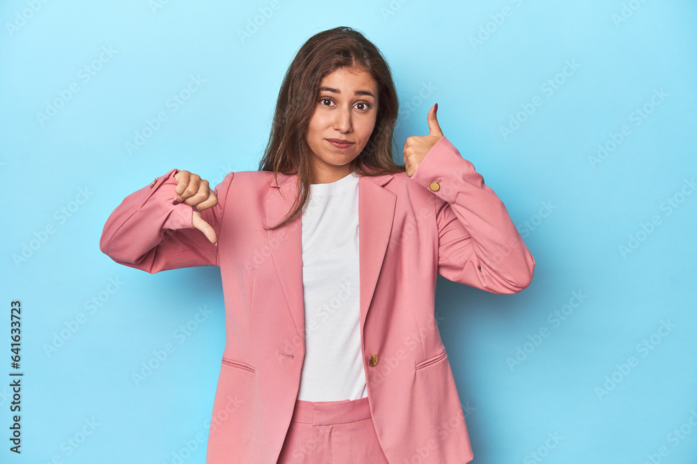 Teen girl in chic pink suit on a blue background showing thumbs up and thumbs down, difficult choose concept