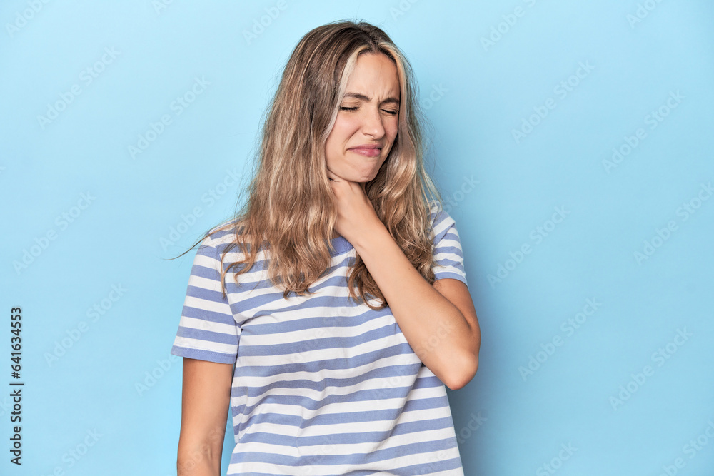 Blonde young caucasian woman in blue studio suffers pain in throat due a virus or infection. Blonde young caucasian woman in blue studio suffers pain in throat due a virus or infection.