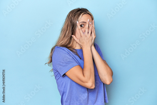 Blonde young caucasian woman in blue studio blink through fingers frightened and nervous.