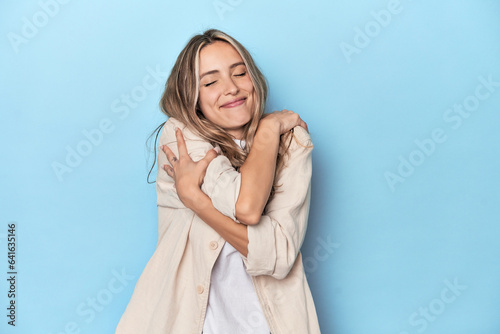 Blonde young caucasian woman in blue studio hugs, smiling carefree and happy.