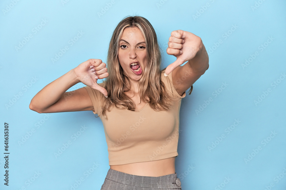 Blonde young caucasian woman in blue studio showing thumb down and expressing dislike. Blonde young caucasian woman in blue studio showing thumb down and expressing dislike.