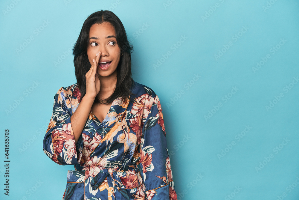 Fototapeta premium Filipina in floral kimono on blue studio is saying a secret hot braking news and looking aside