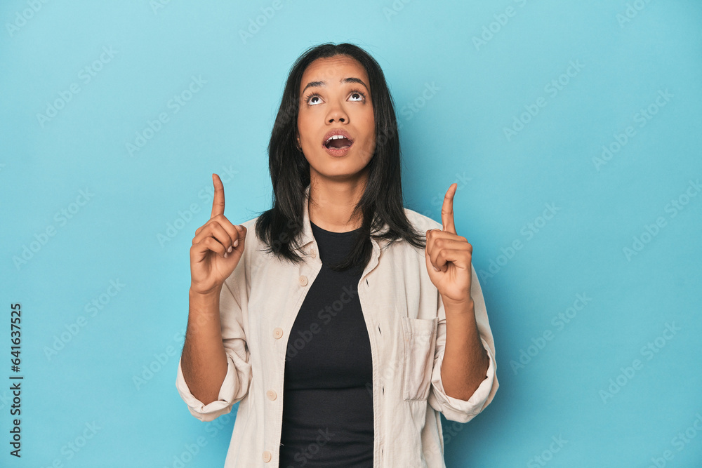 Filipina young woman on blue studio pointing upside with opened mouth. Filipina young woman on blue studio pointing upside with opened mouth.