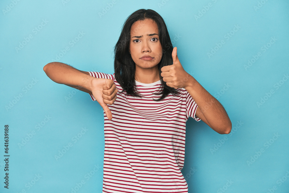 Filipina young woman on blue studio showing thumbs up and thumbs down, difficult choose concept