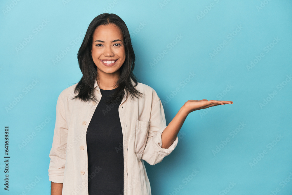 Filipina young woman on blue studio showing a copy space on a palm and holding another hand on waist. Filipina young woman on blue studio showing a copy space on a palm and holding another hand on waist.
