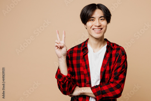 Young smiling happy cheerful man of Asian ethnicity he wear red checkered shirt casual clothes look camera show victory sign isolated on plain pastel light beige background studio. Lifestyle concept. photo