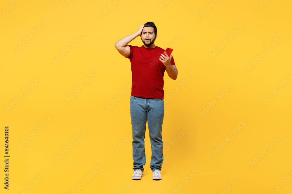 Full body shocked scared young happy Indian man he wearing red t-shirt casual clothes hold head use mobile cell phone look camera isolated on plain yellow orange background studio. Lifestyle concept. Full body shocked scared young happy Indian man he wearing red t-shirt casual clothes hold head use mobile cell phone look camera isolated on plain yellow orange background studio. Lifestyle concept.