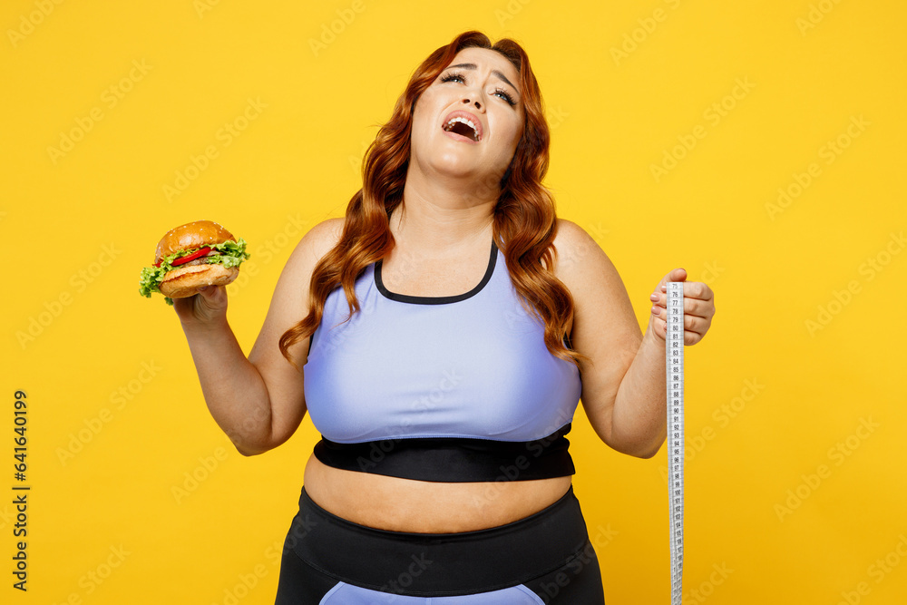 Young tired chubby overweight plus size big fat fit woman wearing blue top warm up training hold measurement tape eat burger isolated on plain yellow background studio home gym. Workout sport concept. Young tired chubby overweight plus size big fat fit woman wearing blue top warm up training hold measurement tape eat burger isolated on plain yellow background studio home gym. Workout sport concept.