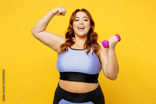 Young fun chubby overweight plus size big fat fit woman wear blue top warm up training hold dumbbells show muscles on hand isolated on plain yellow background studio home gym. Workout sport concept.