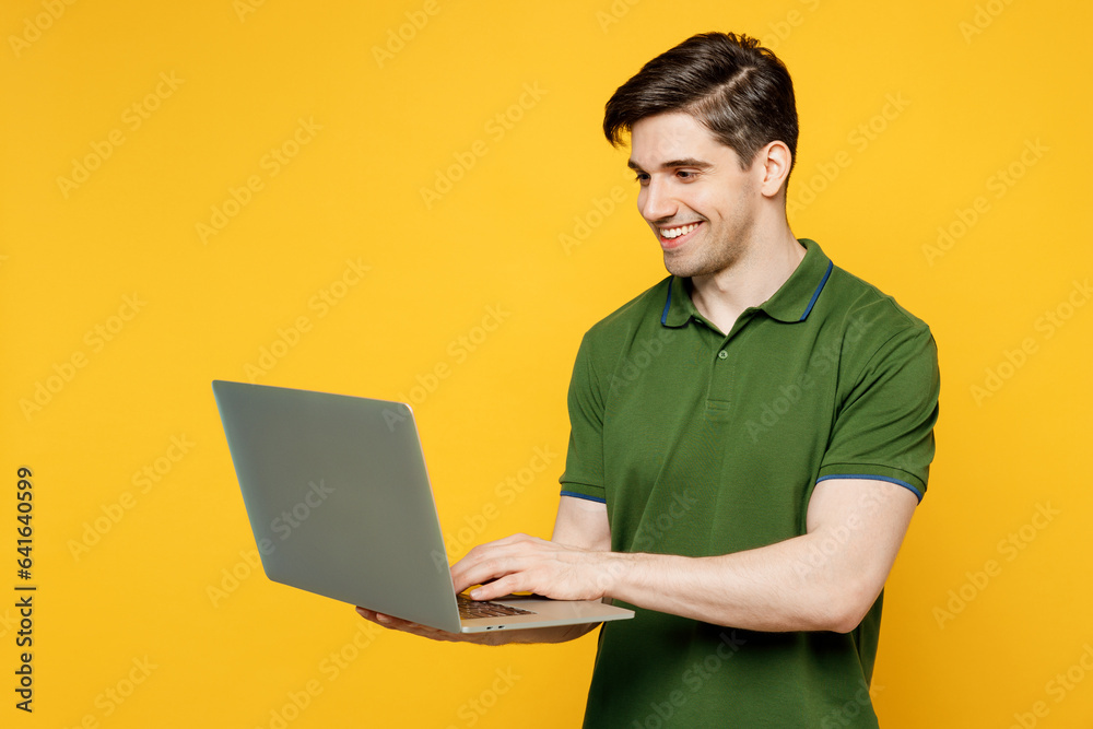Side view young caucasian smiling happy cheerful IT man he wears green t-shirt casual clothes hold use work on laptop pc computer isolated on plain yellow background studio portrait Lifestyle concept Side view young caucasian smiling happy cheerful IT man he wears green t-shirt casual clothes hold use work on laptop pc computer isolated on plain yellow background studio portrait Lifestyle concept