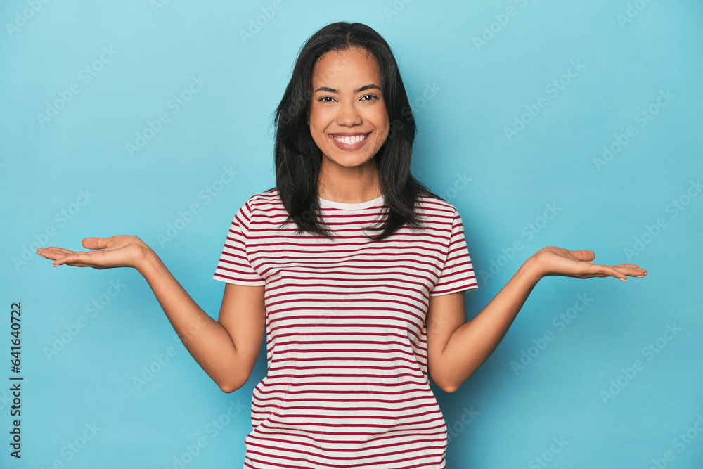 Filipina young woman on blue studio makes scale with arms, feels happy and confident. Filipina young woman on blue studio makes scale with arms, feels happy and confident.