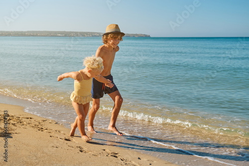 Happy smiling boy holding hand of little girl on beach and runni photo