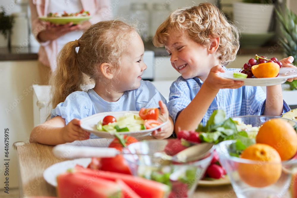 Two happy siblings eating vegetables and fruits in kitchen