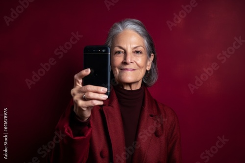 Medium shot portrait photography of a glad mature woman taking a selfie with his mobile against a rich maroon background. With generative AI technology
