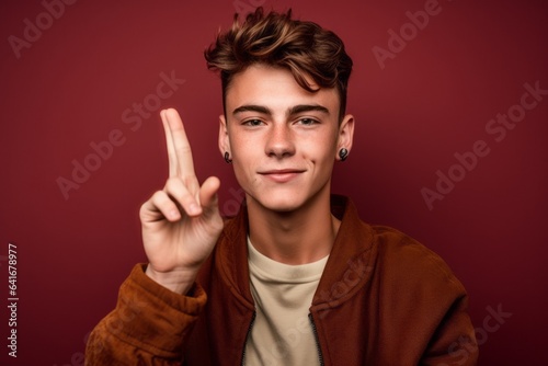 Close-up portrait photography of a beautiful boy in his 20s making a peace and love gesture with the fingers against a rich maroon background. With generative AI technology
