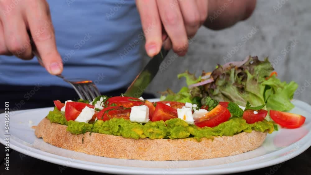 Man has full-fledged nutrition at breakfast with avocado toast. Eat avocado toast to have full-fledged nutrition. Close-up cutting delicious bread toast tomatoes with mashed avocado spread and cheese.