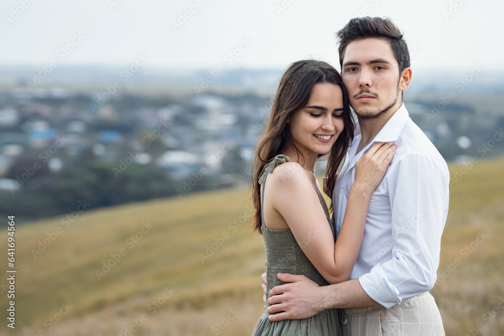 Fototapeta premium Courageous man and positive woman pose standing on hill slope top overlooking large village in cloudy weather. Concept of harmony in relationships and love