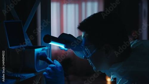 A male expert criminologist removes fingerprints from a jewelry box with a brush. A man gathers evidence using an ultraviolet lamp at a crime scene in a dark apartment lit by red, blue police sirens.