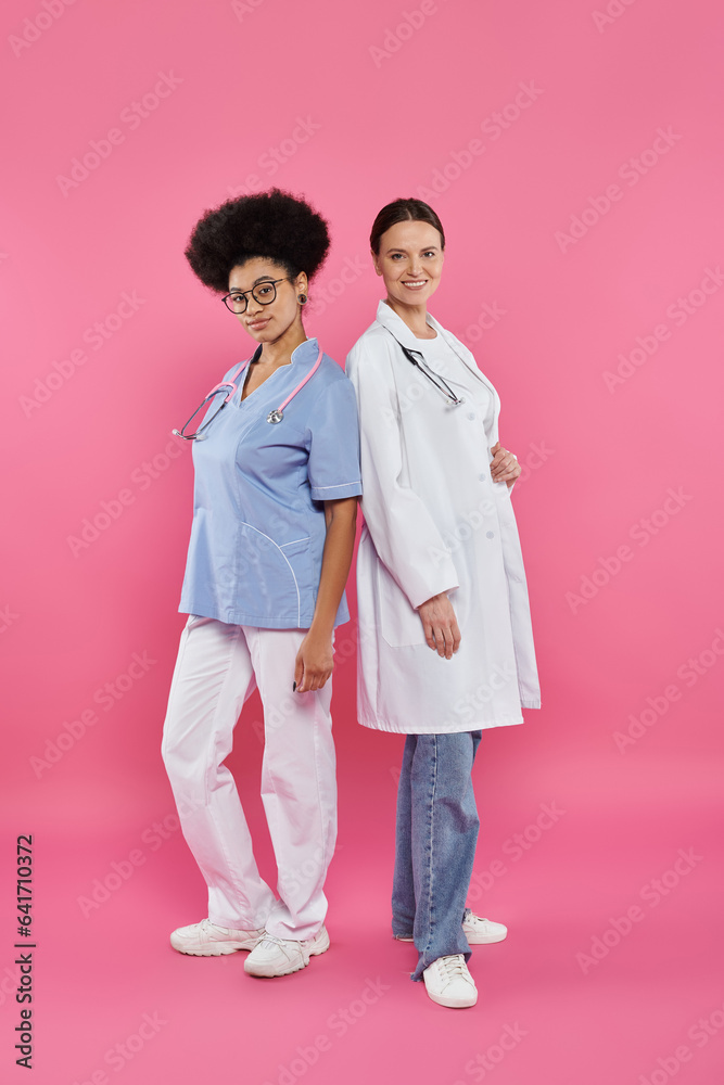 cheerful interracial doctors looking at camera on pink, breast cancer awareness concept cheerful interracial doctors looking at camera on pink, breast cancer awareness concept