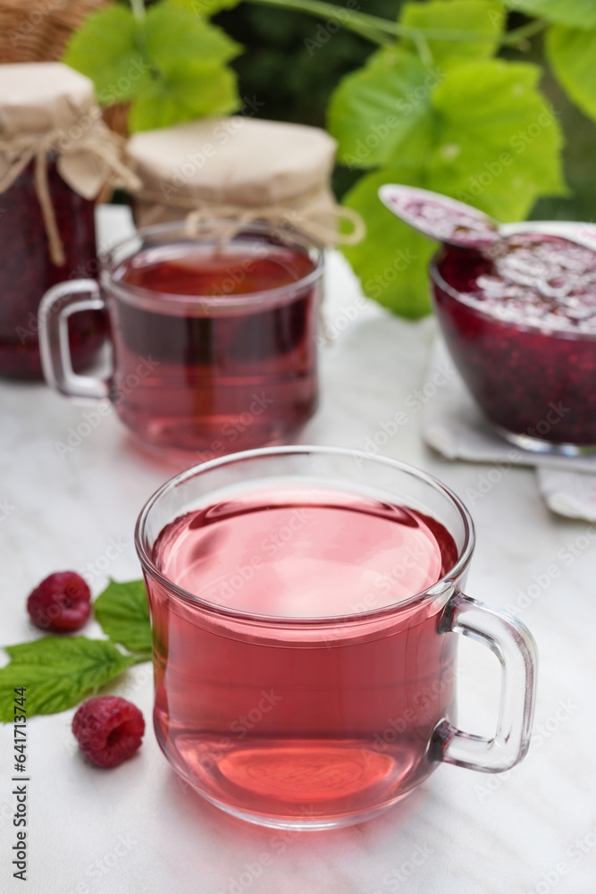 Hot raspberry tea in two transparent cups on a light table. Fresh berries, leaves and jars of jam. Side view, place for text copy space. Hot raspberry tea in two transparent cups on a light table. Fresh berries, leaves and jars of jam. Side view, place for text copy space.
