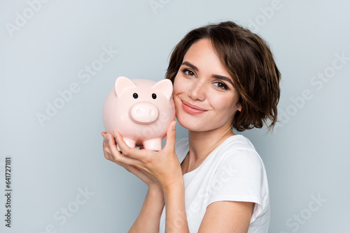 Portrait of good mood girl with bob hairdo wear stylish t-shirt hold pig money box near cheekbone isolated on gray color background