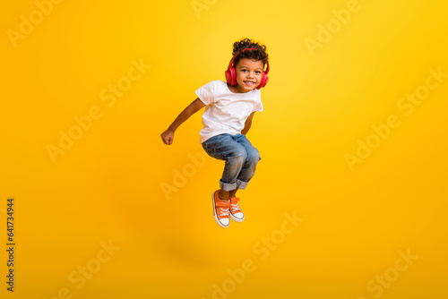Full length photo of carefree excited small boy wear white t-shirt jumping high listening music headphones isolated yellow color background © deagreez