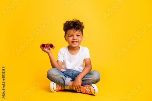 Full length photo of sweet cheerful small boy wear white t-shirt rising binoculars isolated yellow color background