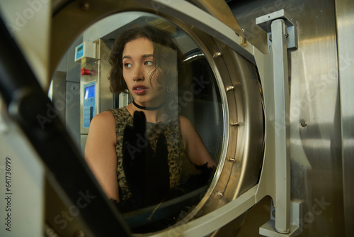trendy young asian woman posing near door of washing machine in public laundry
