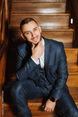 elegant groom with a beard in a beautiful blue jacket and white shirt. Businessman in classic clothes. Brunette man sits on wooden stairs. Studio portrait © Vasil