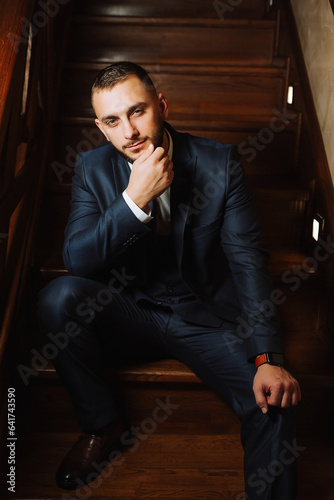 elegant groom with a beard in a beautiful blue jacket and white shirt. Businessman in classic clothes. Brunette man sits on wooden stairs. Studio portrait