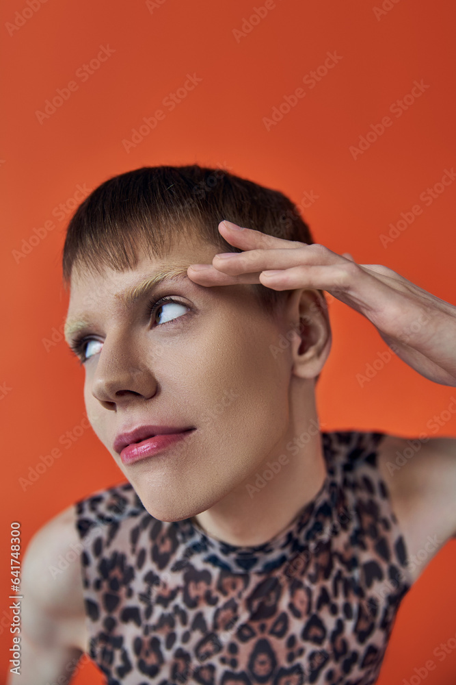 nonbinary person smiling and looking away on orange backdrop, touching eyebrow, queer fashion nonbinary person smiling and looking away on orange backdrop, touching eyebrow, queer fashion
