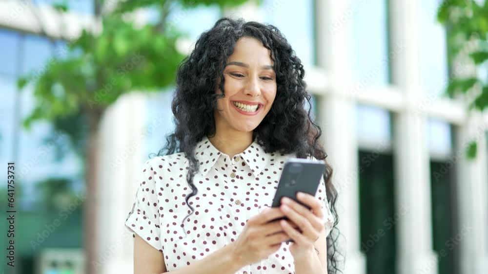 Young smiling female employee is using a smartphone standing on the street near an office building. Happy brunette girl checking email, chatting online, texting with a friend, browsing social media Young smiling female employee is using a smartphone standing on the street near an office building. Happy brunette girl checking email, chatting online, texting with a friend, browsing social media