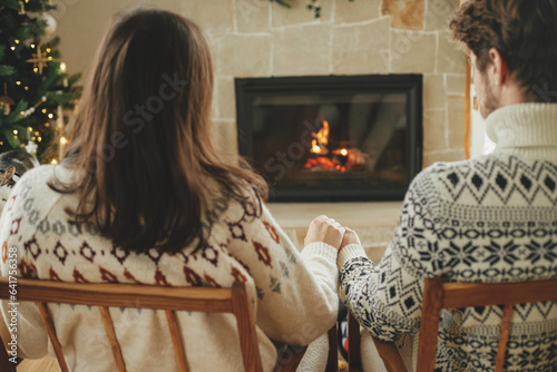 Stylish couple in cozy sweaters relaxing and looking at fireplace with festive mantle on background of stylish decorated christmas tree with lights. Happy young family enjoying winter holidays