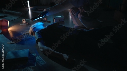 The forensic expert leans over the woman's body and uses an ultraviolet lamp to look for traces and DNA samples. A criminologist in protective overalls and with a work suitcase works at a crime scene. © kinomaster
