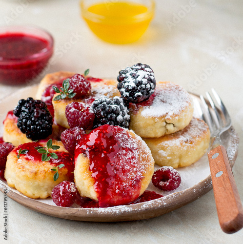 Syrniki serving in a ceramic plate, with raspberries and blackberries, sprinkled with honey and raspberry jam, sprinkled with powdered sugar, horizontal.