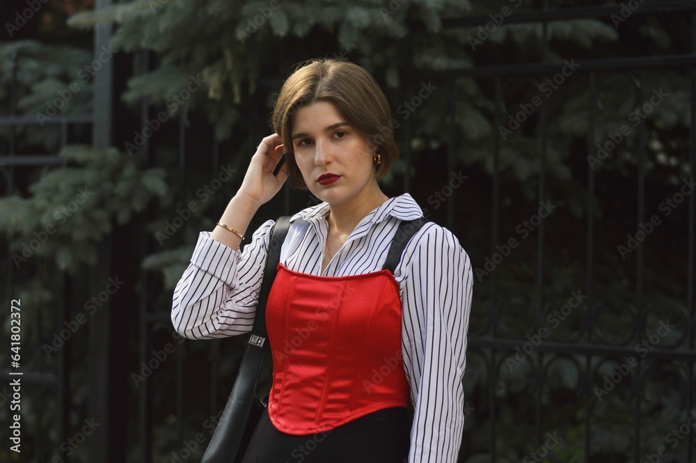 Fototapeta premium A young girl in a fashionable white shirt, red top and black skirt stands near a wall entwined with green leaves and straightens her hair.