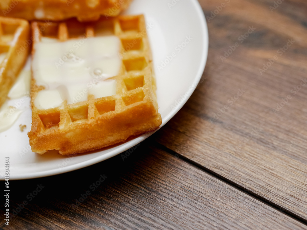 Tasty, golden Belgian waffles covered with sweet condensed milk and served on a white plate on wooden table. Appetizing desert for tea of coffee. Bakery product.