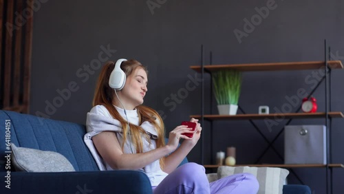 Overjoyed muslim female listening music in headphones and dancing, networking on phone on couch at home. Girl Carefree with smartphone Listening to Music in modern Headphones. People lifestyle concept