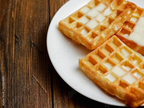 Tasty, golden Belgian waffles covered with sweet condensed milk and served on a white plate on wooden table. Appetizing desert for tea of coffee. Bakery product.