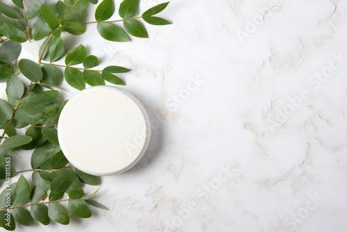 white jar of cream on a white marble background with green leaves scattered around it