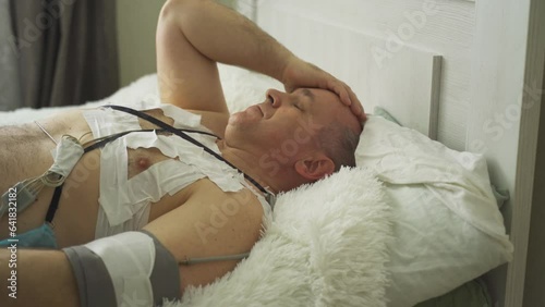 A man with a holter installed reveals malfunctions in the cardiovascular system. Lying on the bed, holding his head, rubbing his eyes. photo
