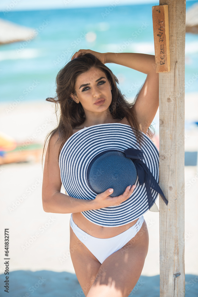 Beautiful brunette girl in a white bathing suit.Portrait of professional swimwear model posing provocatively outdoor.Smiling sensual lady with long legs posing in white bikini and hat the beach.