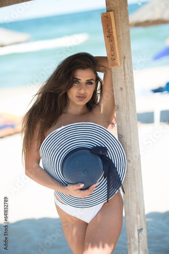 Obraz na płótnie Beautiful brunette girl in a white bathing suit.Portrait of professional swimwear model posing provocatively outdoor.Smiling sensual lady with long legs posing in white bikini and hat the beach. w salonie